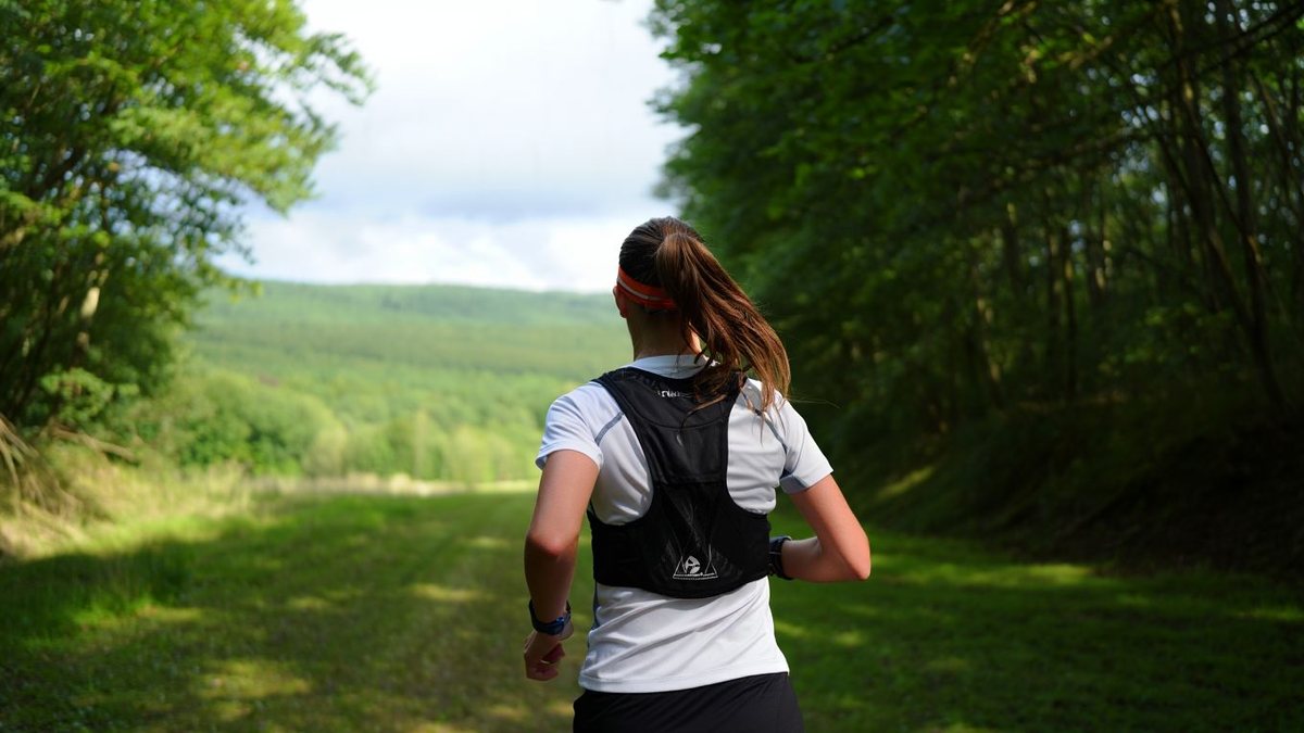 Coureur amateur sur sentier forestier vallonné avec sac de trail discret sans texte ni marque visible
